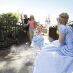 Une jeune fille en costume de princesse court avec enthousiasme vers une femme habillée en Cendrillon en robe bleue à l'Auberge de Cendrillon, avec sa famille souriant derrière elle sous le soleil