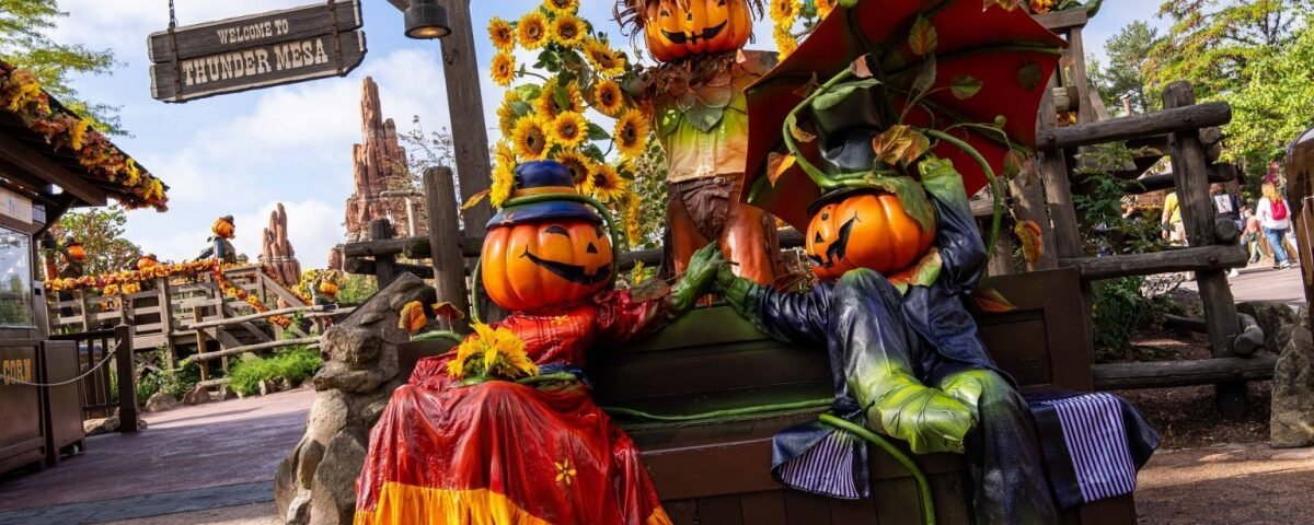 Deux personnages à tête de citrouille vêtus de costumes colorés de Disneyland Paris sont assis sur un banc de parc entouré de tournesols, avec un panneau « Thunder mesa » en arrière-plan, célébrant Halloween.