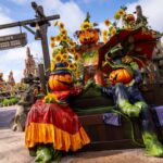 Deux personnages à tête de citrouille vêtus de costumes colorés de Disneyland Paris sont assis sur un banc de parc entouré de tournesols, avec un panneau « Thunder mesa » en arrière-plan, célébrant Halloween.