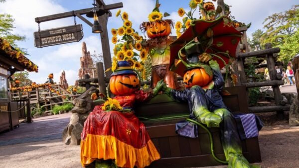 Deux personnages à tête de citrouille vêtus de costumes colorés de Disneyland Paris sont assis sur un banc de parc entouré de tournesols, avec un panneau « Thunder mesa » en arrière-plan, célébrant Halloween.
