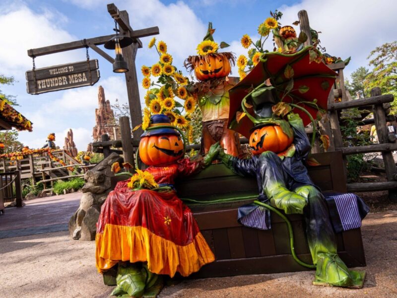 Deux personnages à tête de citrouille vêtus de costumes colorés de Disneyland Paris sont assis sur un banc de parc entouré de tournesols, avec un panneau « Thunder mesa » en arrière-plan, célébrant Halloween.