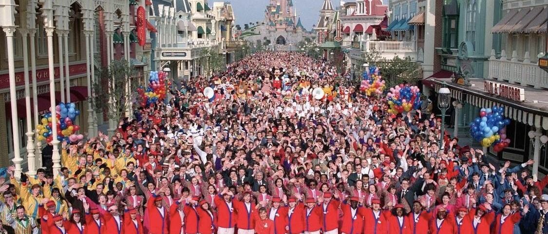Une grande foule s'est rassemblée sur Main Street à Disneyland Paris, avec des personnes tenant des ballons et des personnages Disney visibles. La première rangée comprend des acteurs en tenue de cérémonie rouge et blanche.