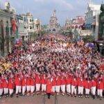 Une grande foule s'est rassemblée sur Main Street à Disneyland Paris, avec des personnes tenant des ballons et des personnages Disney visibles. La première rangée comprend des acteurs en tenue de cérémonie rouge et blanche.