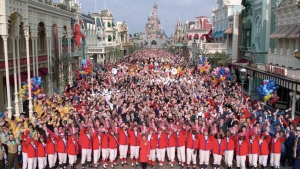 Une grande foule s'est rassemblée sur Main Street à Disneyland Paris, avec des personnes tenant des ballons et des personnages Disney visibles. La première rangée comprend des acteurs en tenue de cérémonie rouge et blanche.