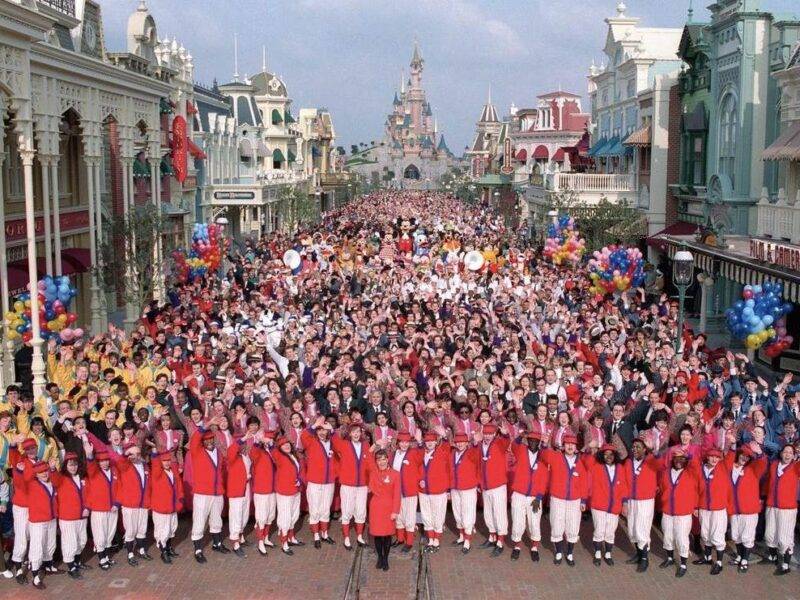 Une grande foule s'est rassemblée sur Main Street à Disneyland Paris, avec des personnes tenant des ballons et des personnages Disney visibles. La première rangée comprend des acteurs en tenue de cérémonie rouge et blanche.
