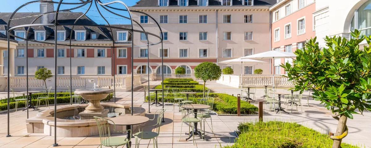 Cour d'un hôtel de luxe avec une élégante pergola en métal, des sièges extérieurs, une fontaine centrale et des haies bien entretenues, dans le contexte de Disneyland Paris.