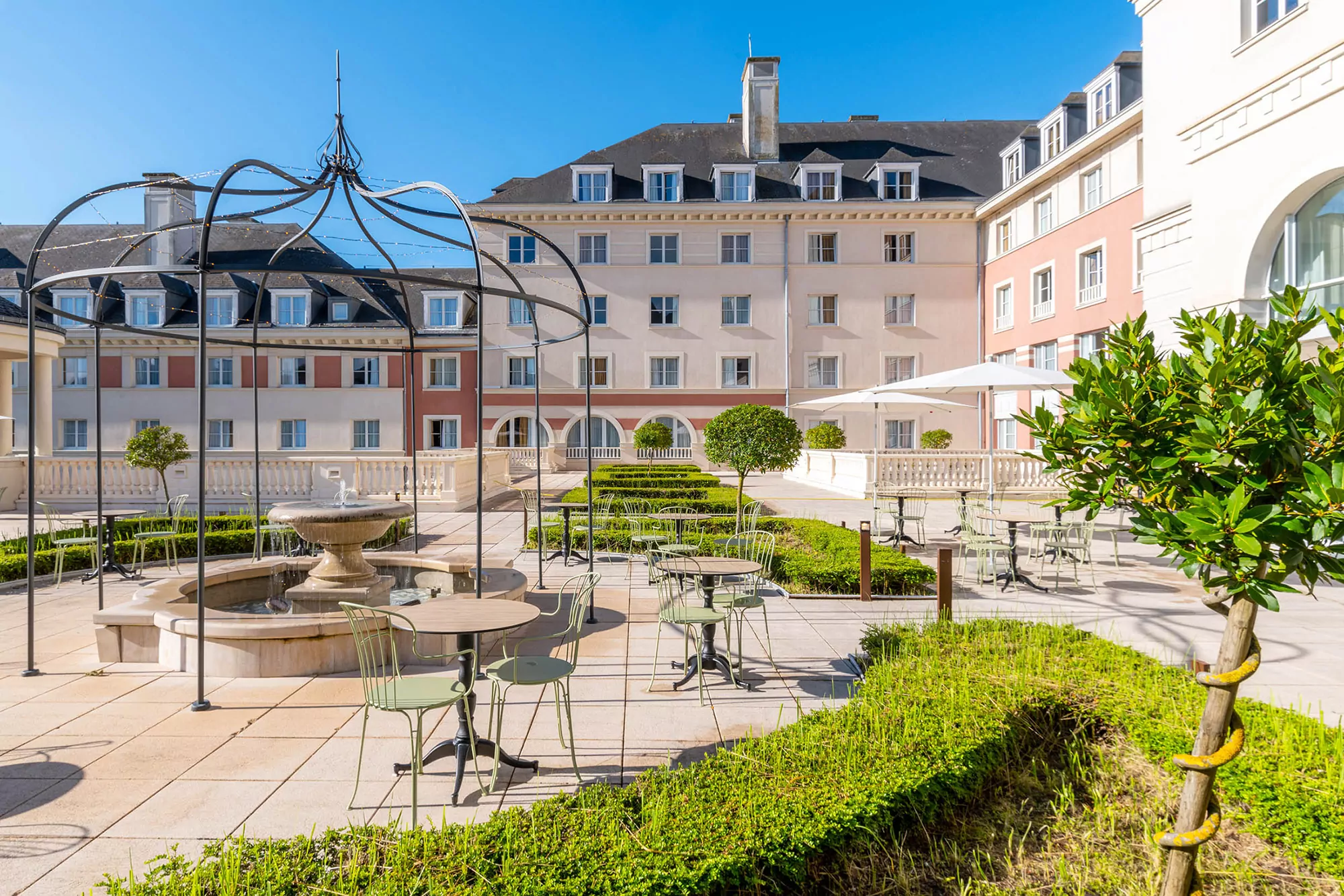 Cour d'un hôtel de luxe avec une élégante pergola en métal, des sièges extérieurs, une fontaine centrale et des haies bien entretenues, dans le contexte de Disneyland Paris.