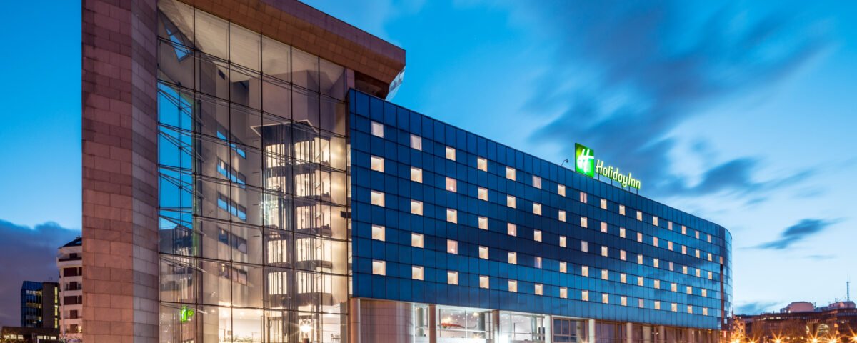 Bâtiment hôtelier moderne éclairé au crépuscule avec une façade en verre, arborant le logo Disneyland Paris, situé au bord d'un plan d'eau calme avec un ciel flou indiquant une longue exposition.