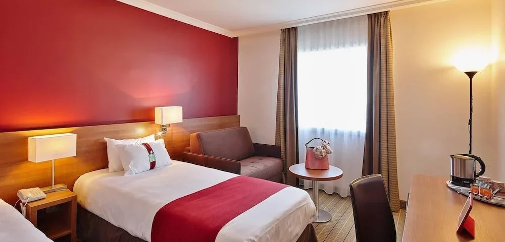 Une chambre d'hôtel Holiday Inn bien aménagée avec un grand lit doté d'un jeté rouge, un mur d'accent rouge vif, un coin bureau et des sièges moelleux près d'une fenêtre drapée de rideaux transparents.