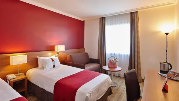 Une chambre d'hôtel Holiday Inn bien aménagée avec un grand lit doté d'un jeté rouge, un mur d'accent rouge vif, un coin bureau et des sièges moelleux près d'une fenêtre drapée de rideaux transparents.