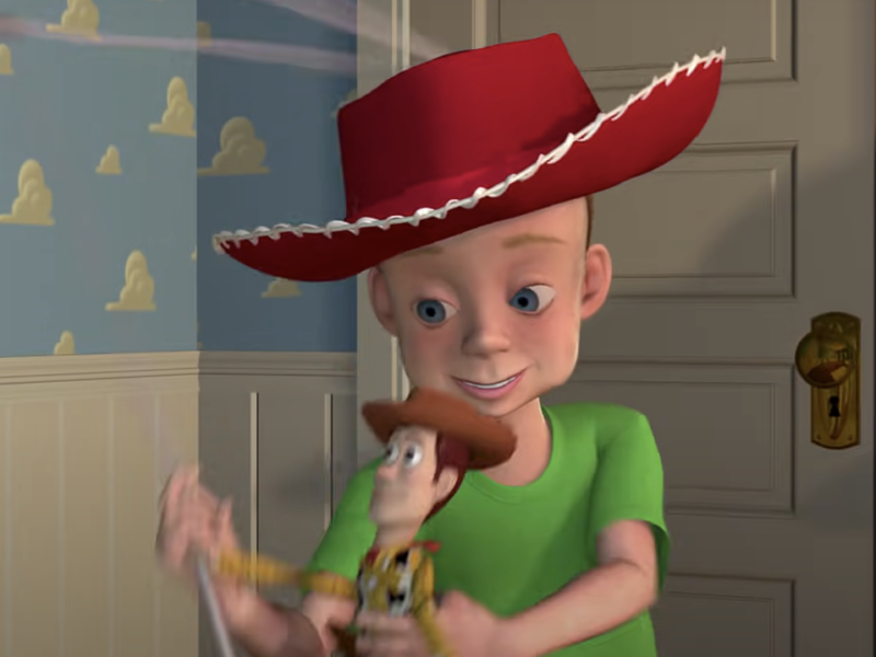 Un jeune garçon au sourire éclatant, coiffé d'un grand chapeau de cowboy rouge et d'un t-shirt vert, tenant joyeusement un jouet Woody du film Toy Story de Pixar dans une pièce au papier peint à motifs de nuages.