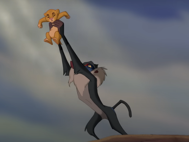 Une scène de "Le Roi Lion" montrant Rafiki tenant Simba en l'air sur Pride Rock sous un ciel dramatique.