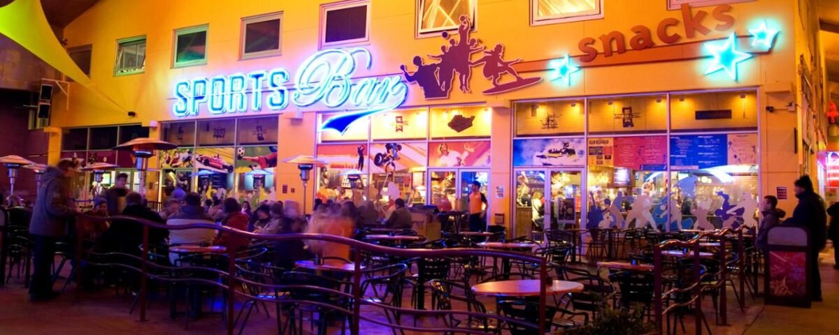 Un bar sportif animé dans Disney Village avec des enseignes au néon, animé de gens assis à des tables et discutant, avec des comptoirs servant des collations en arrière-plan.