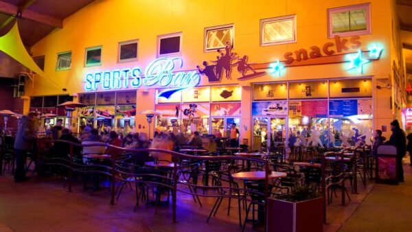 Un bar sportif animé dans Disney Village avec des enseignes au néon, animé de gens assis à des tables et discutant, avec des comptoirs servant des collations en arrière-plan.