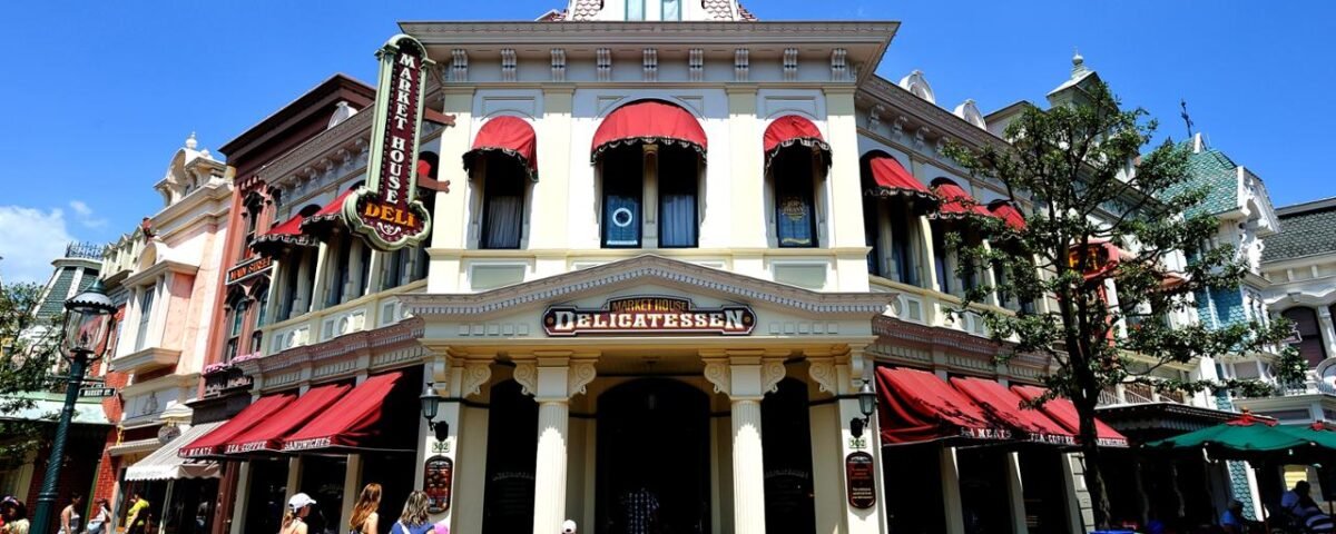 Une scène de rue animée à Disneyland Paris, avec un bâtiment de deux étages avec des auvents rouges, étiqueté Market House Deli et hôtel, sous un ciel bleu clair, avec des gens marchant le long du trottoir.