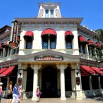 Une scène de rue animée à Disneyland Paris, avec un bâtiment de deux étages avec des auvents rouges, étiqueté Market House Deli et hôtel, sous un ciel bleu clair, avec des gens marchant le long du trottoir.