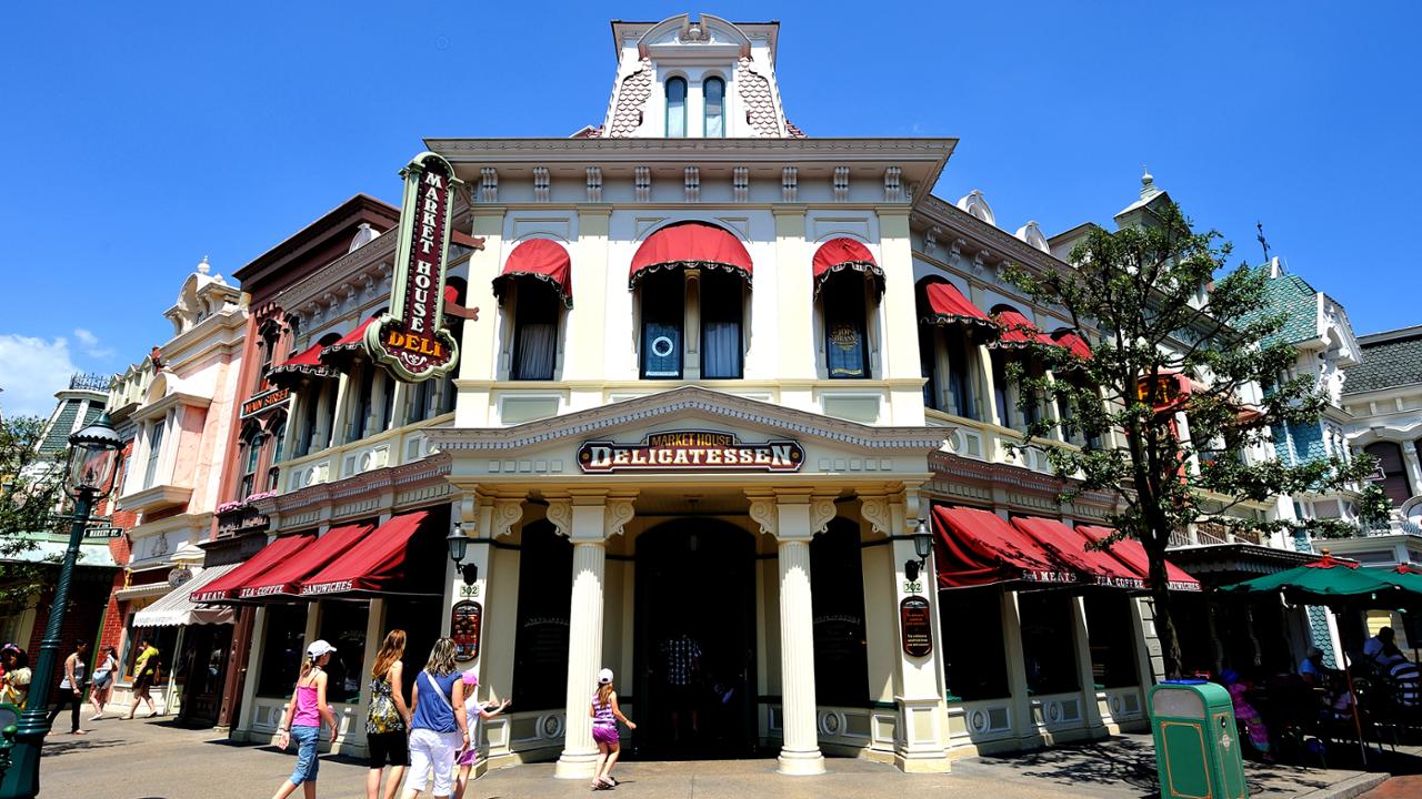 Une scène de rue animée à Disneyland Paris, avec un bâtiment de deux étages avec des auvents rouges, étiqueté Market House Deli et hôtel, sous un ciel bleu clair, avec des gens marchant le long du trottoir.