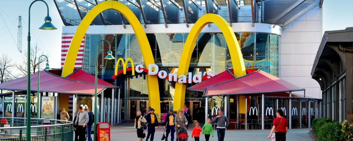 Extérieur du restaurant McDonald's moderne situé près de Disneyland Paris, avec de grandes arches dorées, une façade en verre et des gens se promenant, capturés à la lumière du jour.
