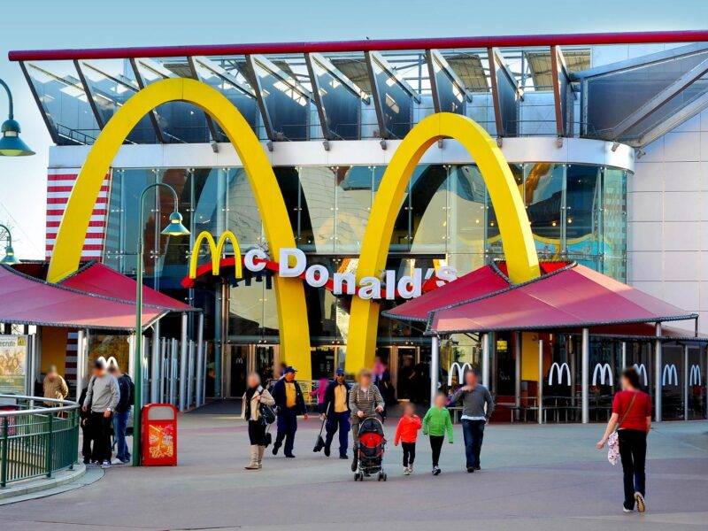 Extérieur du restaurant McDonald's moderne situé près de Disneyland Paris, avec de grandes arches dorées, une façade en verre et des gens se promenant, capturés à la lumière du jour.