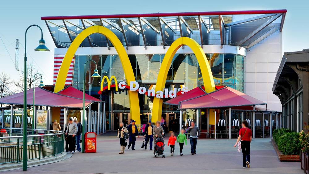 Extérieur du restaurant McDonald's moderne situé près de Disneyland Paris, avec de grandes arches dorées, une façade en verre et des gens se promenant, capturés à la lumière du jour.