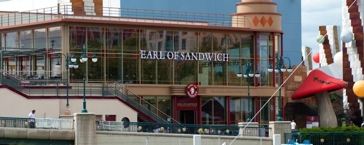 Vue extérieure du restaurant "Earl of Sandwich" situé à Disneyland Paris, avec un plan d'eau au premier plan et des éléments de décoration autour de son architecture moderne.