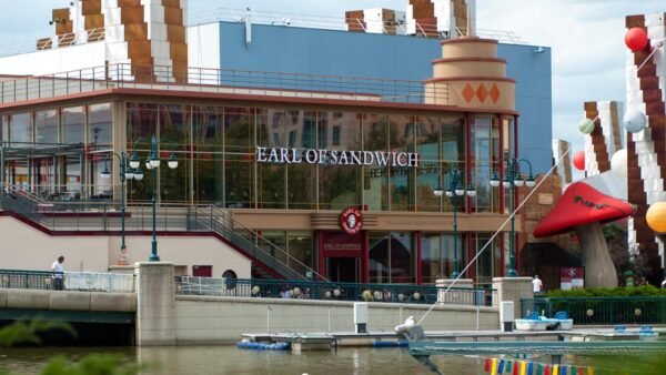 Vue extérieure du restaurant "Earl of Sandwich" situé à Disneyland Paris, avec un plan d'eau au premier plan et des éléments de décoration autour de son architecture moderne.