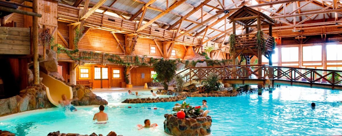 Un parc aquatique intérieur animé avec des poutres et des structures en bois, comprenant une piscine, des toboggans et plusieurs personnes profitant de l'eau. Les piscines brillent sous un éclairage naturel éclatant qui illumine la scène.