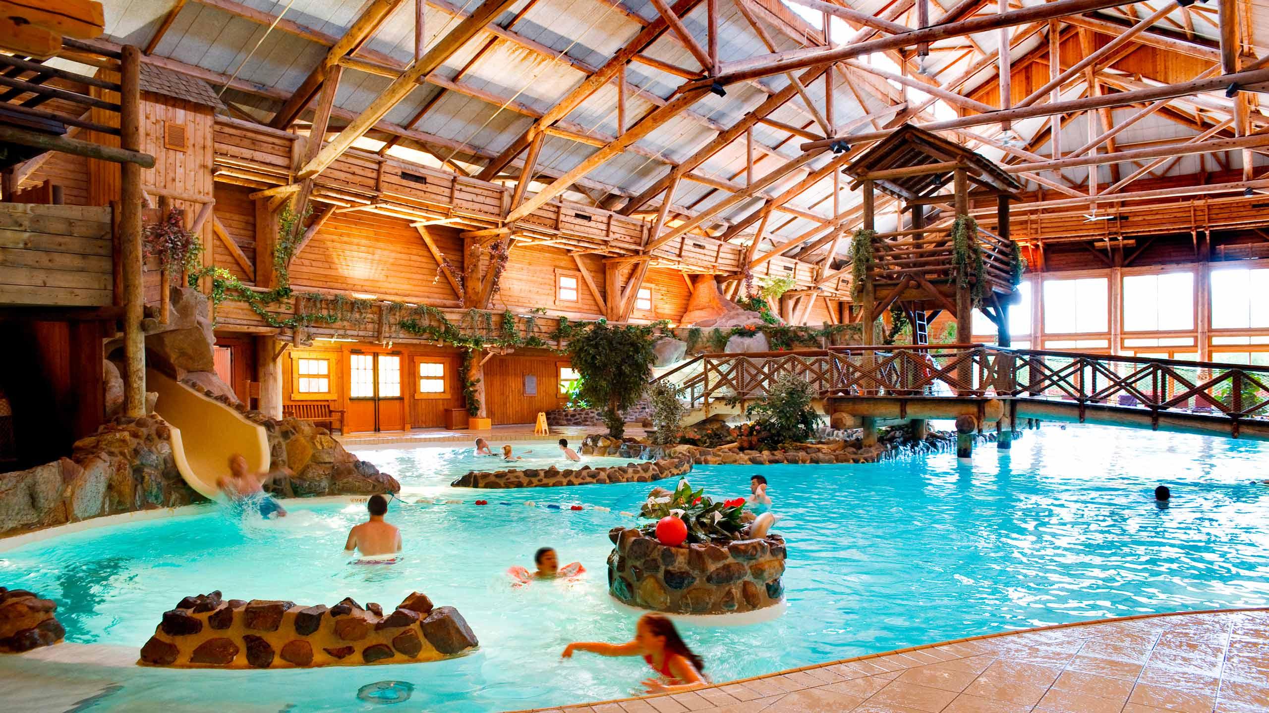 Un parc aquatique intérieur animé avec des poutres et des structures en bois, comprenant une piscine, des toboggans et plusieurs personnes profitant de l'eau. Les piscines brillent sous un éclairage naturel éclatant qui illumine la scène.