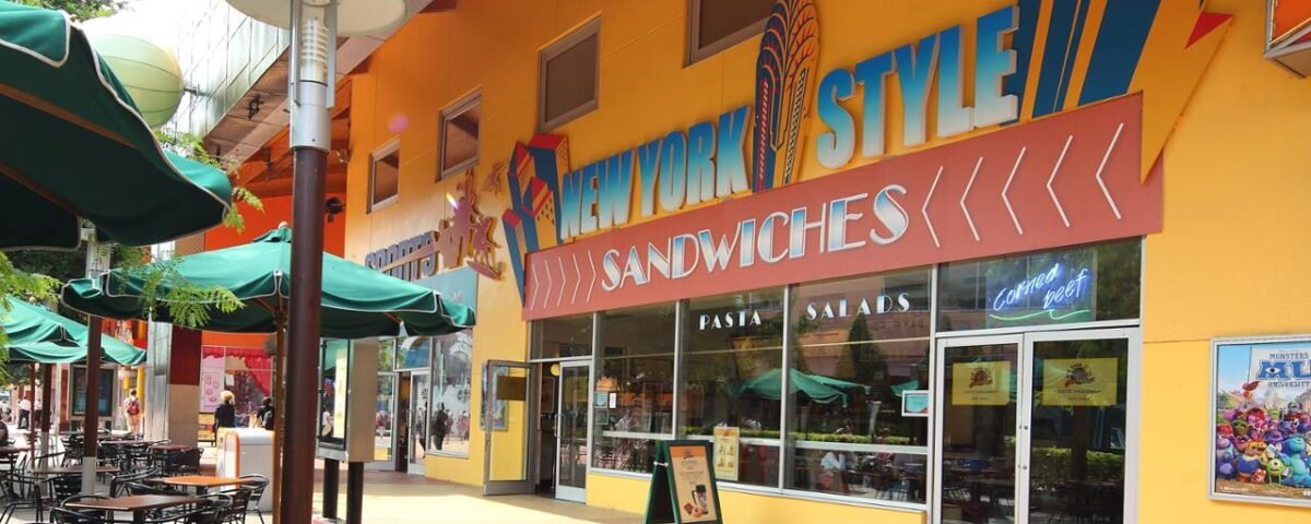 Une façade de restaurant colorée avec une pancarte « sandwichs de style new-yorkais », avec des sièges à l'extérieur sous des parasols verts dans une zone piétonne ensoleillée de Disney Village.