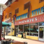 Une façade de restaurant colorée avec une pancarte « sandwichs de style new-yorkais », avec des sièges à l'extérieur sous des parasols verts dans une zone piétonne ensoleillée de Disney Village.