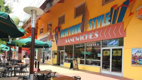 Une façade de restaurant colorée avec une pancarte « sandwichs de style new-yorkais », avec des sièges à l'extérieur sous des parasols verts dans une zone piétonne ensoleillée de Disney Village.