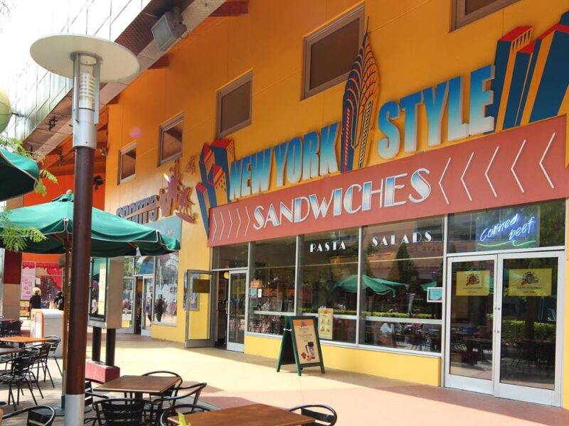 Une façade de restaurant colorée avec une pancarte « sandwichs de style new-yorkais », avec des sièges à l'extérieur sous des parasols verts dans une zone piétonne ensoleillée de Disney Village.