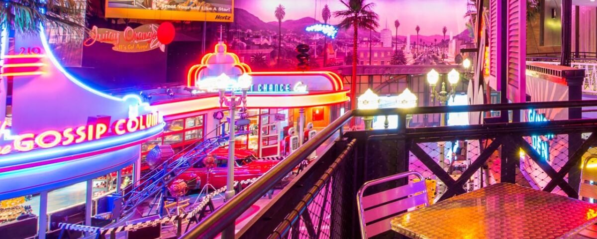 Une scène intérieure vibrante à Disneyland Paris avec des enseignes au néon, dont une qui indique "Gossip Grill", avec en toile de fond un coucher de soleil et un escalier métallique au premier plan dans la zone du Restaurant En Coulisse.