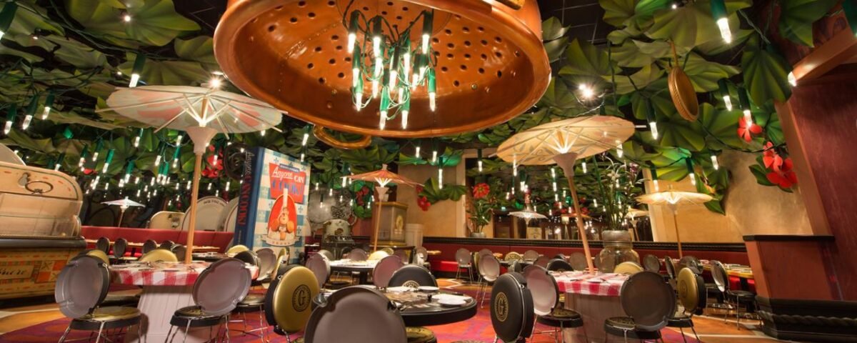 Intérieur d'un restaurant fantaisiste à Disneyland Paris, style Bistrot Chez Rémy, avec des décorations surdimensionnées de feuilles et de passoires au-dessus, et des sièges colorés et éclectiques disposés autour de tables circulaires.