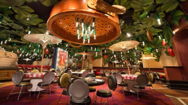 Intérieur d'un restaurant fantaisiste à Disneyland Paris, style Bistrot Chez Rémy, avec des décorations surdimensionnées de feuilles et de passoires au-dessus, et des sièges colorés et éclectiques disposés autour de tables circulaires.