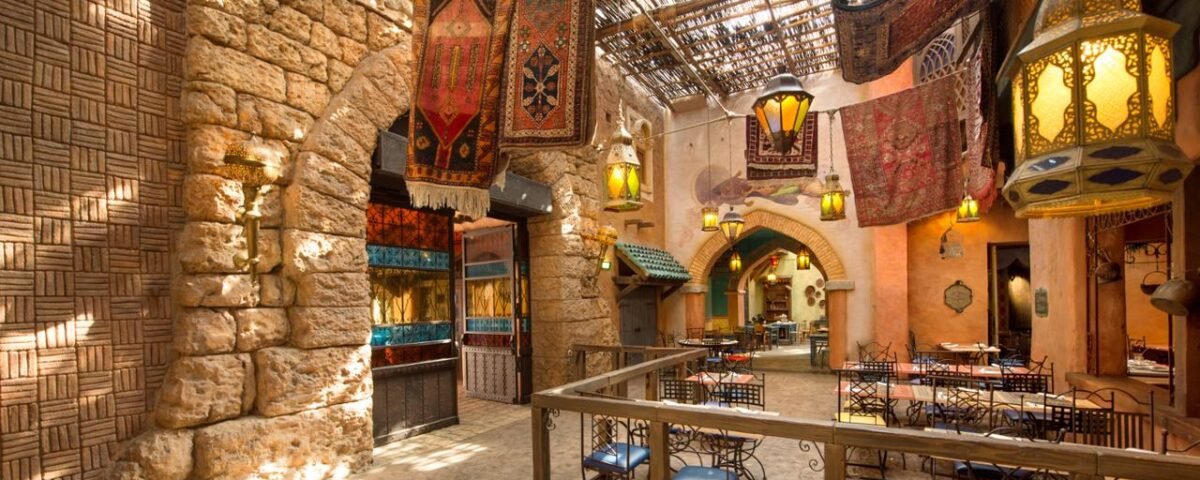 Restaurant intérieur sur le thème de l'Agrabah Café avec lanternes suspendues, tapis colorés, murs en pierre et toit en osier. La salle à manger comporte des tables vides avec des chaises bleues.