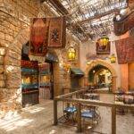 Restaurant intérieur sur le thème de l'Agrabah Café avec lanternes suspendues, tapis colorés, murs en pierre et toit en osier. La salle à manger comporte des tables vides avec des chaises bleues.