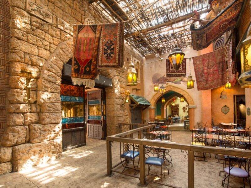 Restaurant intérieur sur le thème de l'Agrabah Café avec lanternes suspendues, tapis colorés, murs en pierre et toit en osier. La salle à manger comporte des tables vides avec des chaises bleues.