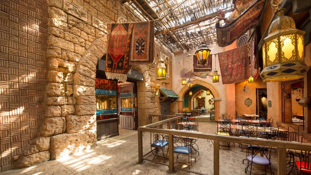 Restaurant intérieur sur le thème de l'Agrabah Café avec lanternes suspendues, tapis colorés, murs en pierre et toit en osier. La salle à manger comporte des tables vides avec des chaises bleues.