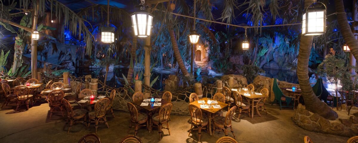 Intérieur d'un restaurant à thème ressemblant à une jungle luxuriante avec des parois rocheuses, des plantes tropicales et des cimes d'arbres. Des tables et des chaises en bois sont disposées partout, éclairées par un éclairage ambiant doux inspiré du capitaine Jack.