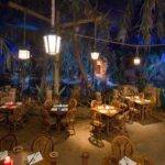 Intérieur d'un restaurant à thème ressemblant à une jungle luxuriante avec des parois rocheuses, des plantes tropicales et des cimes d'arbres. Des tables et des chaises en bois sont disposées partout, éclairées par un éclairage ambiant doux inspiré du capitaine Jack.