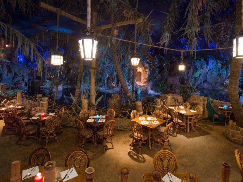 Intérieur d'un restaurant à thème ressemblant à une jungle luxuriante avec des parois rocheuses, des plantes tropicales et des cimes d'arbres. Des tables et des chaises en bois sont disposées partout, éclairées par un éclairage ambiant doux inspiré du capitaine Jack.