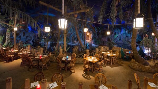 Intérieur d'un restaurant à thème ressemblant à une jungle luxuriante avec des parois rocheuses, des plantes tropicales et des cimes d'arbres. Des tables et des chaises en bois sont disposées partout, éclairées par un éclairage ambiant doux inspiré du capitaine Jack.