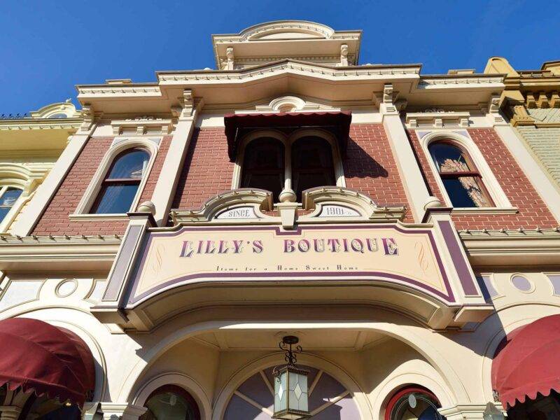 Une image de la façade de la boutique Lilly, présentant une élégante architecture victorienne avec une palette de couleurs pastel. Le panneau et les détails ornés sont mis en valeur sous un ciel bleu clair.