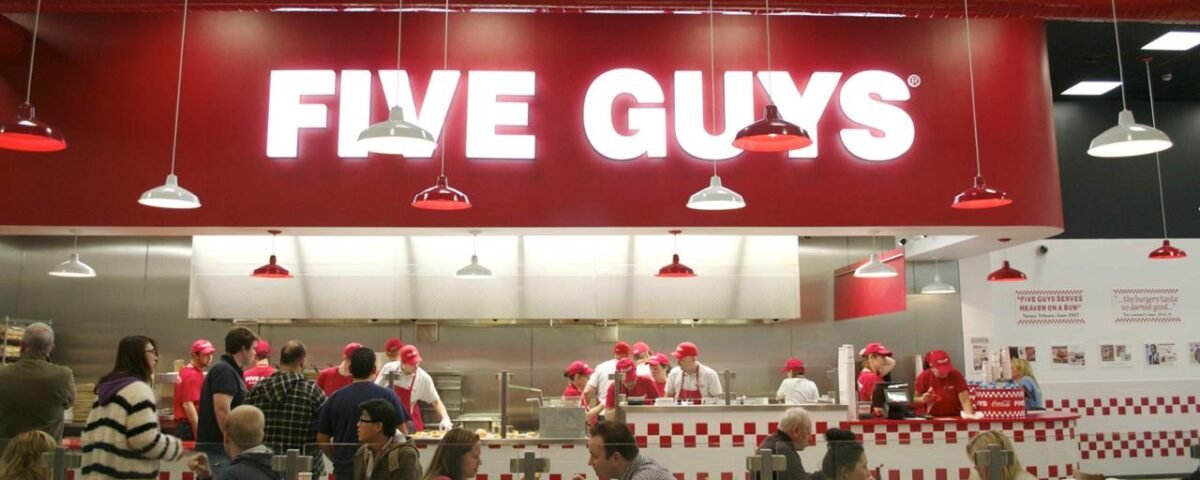 Intérieur d'un restaurant Five Guys à Disneyland Paris avec des clients au comptoir, du personnel en chapeaux rouges préparant la nourriture et un panneau rouge audacieux au-dessus. Thème rouge et blanc avec suspensions et tableaux de menu visibles