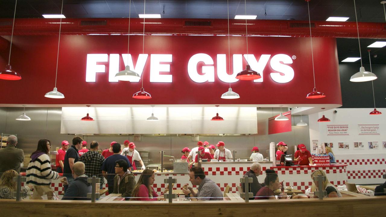 Intérieur d'un restaurant Five Guys à Disneyland Paris avec des clients au comptoir, du personnel en chapeaux rouges préparant la nourriture et un panneau rouge audacieux au-dessus. Thème rouge et blanc avec suspensions et tableaux de menu visibles