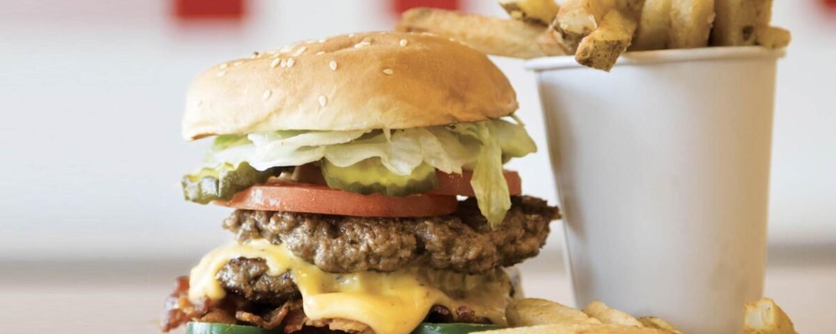Un double cheeseburger avec laitue, tomate et oignons de Five Guys, servi avec un côté de frites dans une tasse blanche, posé sur une table en bois avec un fond rouge et blanc flou.