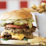 Un double cheeseburger avec laitue, tomate et oignons de Five Guys, servi avec un côté de frites dans une tasse blanche, posé sur une table en bois avec un fond rouge et blanc flou.
