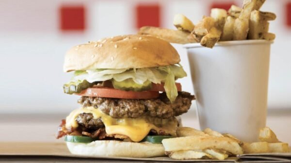 Un double cheeseburger avec laitue, tomate et oignons de Five Guys, servi avec un côté de frites dans une tasse blanche, posé sur une table en bois avec un fond rouge et blanc flou.