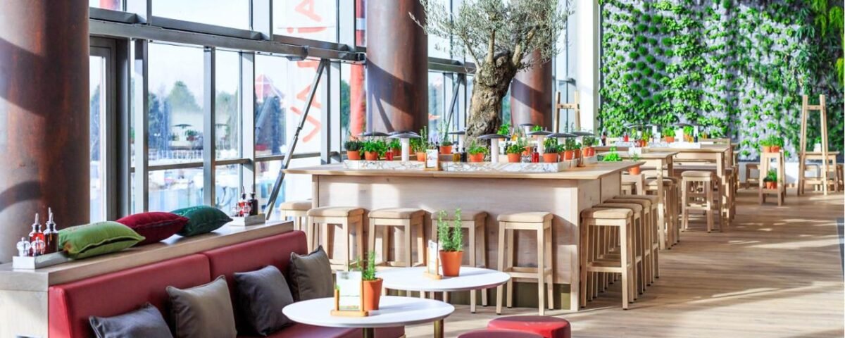 Un intérieur de restaurant moderne avec de grandes fenêtres, un mur végétal intérieur, des tables en bois et un mélange de canapés et de tabourets dans une ambiance vibrante et aérée inspirée de Disneyland Paris.